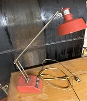 Vintage red adjustable desk lamp with metal arm and matching red shade and base photographed on a wooden surface.