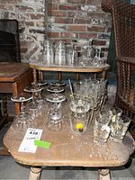 Overview of the lot showing various glassware arranged on and under a wooden table with a rough brick wall background. Visible stemware, tumblers, condiment holder with spoon, and glasses in cart holder.