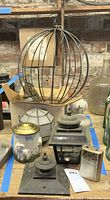 Photo showing a metal spherical cage light fixture, two black metal lantern-style outdoor light fixtures, ceramic container with brass lid, small ceramic/metal component, and other metal lamp parts.