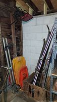 Downhill skis, wood skis, sleds stored in wooden crate and leaning against wall with fishing rods inside crate.