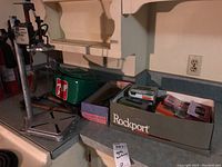 Drill press stand on counter next to soft-sided 7UP cooler bag and a Rockport shoe box containing hardware items and small tool kits
