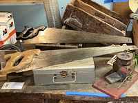 Two vintage hand saws with wooden handles are on a table next to a metal toolbox and a small mechanical scale.