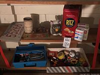 Shelf imaged showing vintage collectibles including playing cards, Furby toy, tins, Band-Aid boxes, blue medical case with stethoscope, boot jack corkscrew, and license plate.