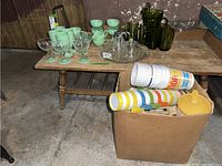 Overview of the lot showing vintage glass bottles, assorted plastic colorful cups in a box, pale green plastic cups and goblets, clear etched glass dessert glasses, and plastic yellow pitcher.
