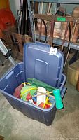 Blue plastic bin open showing assorted outdoor toys including green flippers and rubber shoes, located on a concrete floor with shelving and other items in background