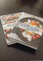 Photo of two hardcover cookbooks laid flat on a dark surface. The top book is 'Jerusalem,' and underneath is 'Plenty.' The covers are clean with vivid food photography.