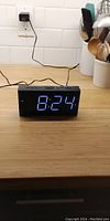 Photo of black digital alarm clock showing the current time 8:24 on bright blue LED display on wooden surface.