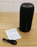 Black MagnaBoom MB-395B Bluetooth speaker standing upright on wooden surface with instruction manual and charging cable beside it. Speaker has a textured grip and speaker grille on top.