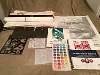 Wide view of the lot contents showing rolled papers, drawing pads, instructional book, stencil sheets, paint pods, and brush holder arranged on a bed.