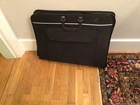 Closed black artist portfolio case standing upright against a wall, showing carrying handles and outer VELCRO pocket.