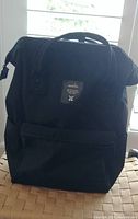 Front view of black Anello backpack with front zippered pocket and top carry handle.
