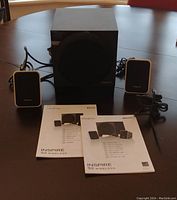 Full view of speaker set including black receiver, pair of white satellite speakers, power cable, and printed manuals on table surface