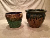 Photo showing two glazed ceramic pots side by side on a beige fabric background, highlighting their colors and embossed designs.