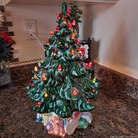 Front view of a green ceramic Christmas tree with multicolored miniature lights embedded in the foliage. Base has ceramic presents, teddy bear, and guitar.