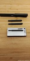 Matte black Lamy Safari fountain pen, two spare black ink cartridges in original Lamy box placed on wooden surface.