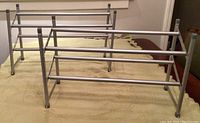 Photo showing two silver metal shoe racks with 2 horizontal tiers each. Expandable width metal bars and vertical legs visible. Beige fabric background.