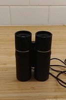 Front view of closed compact black binoculars with eyepiece lenses visible and neck strap.