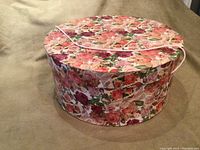 Closed round vintage floral print hat box showing pink, red, and cream roses with a pink cord handle on the lid.