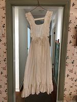 Full view of vintage cream apron hanging on wire hanger showing overall design, length, ruffle detail with straps and hem.