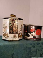 Photo showing both vintage Asian artwork tins, hexagonal and cylindrical, placed on a blue-green patterned cloth.