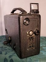 Angled side view of the black textured vintage Cine Kodak Model B movie camera showing the carrying strap and crank handle.