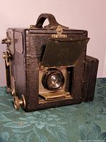 Front angle view of the Graflex Series 23069 camera showing textured leather body, lens with metal cover partially open, and handle on top.