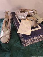 Pair of metallic silver Roberto Capucci ladies shoes shown resting on a box with a receipt. One shoe displayed sideways showing the open toe, ankle strap with buckle, and patterned sides.