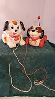Front facing view of two pull along toy dogs, white with black ears and red scarf dog, and a brown and white dog with wheels and antenna