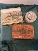 Three vintage tins displayed on green cloth background: scenic castle tin, rust-colored Horse Shoe Cigarette tin, small round decorative tin with woman at sewing machine illustration.