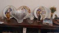 View of the three Knowles collector plates arranged behind crystal bowl, glass jar, candle holder, and other items on a wooden surface.