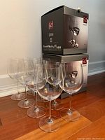 Eight clear crystal wine glasses arranged in two rows next to two original F&D packaging boxes, on a wooden floor.