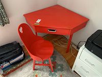 Red painted hardwood corner desk and chair placed in a corner on wooden flooring, showing overall presence and shape.