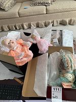 Four boxed baby dolls with pastel dresses and lace bonnets lying on a couch and table. Dolls appear to be in protective cellophane and tissue paper packaging inside their boxes.