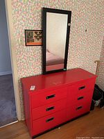 Red painted hardwood dresser with six drawers and black drawer pulls, rectangular black framed mirror leaning on wall above it. Shows visible scuffs and paint wear on top surface.