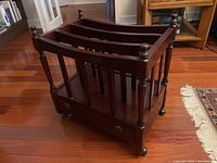 Full view of wooden magazine rack showing vertical slats, rounded posts with finials, metal casters, and drawer on bottom.