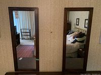 Two matching rectangular mirrors hung vertically on a wall with dark wood frames reflecting bedroom and living area.