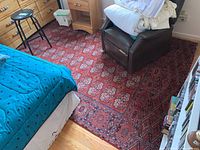 Image from bedroom showing the machine woven red area rug with traditional pattern and cutout near one edge for ventilation. Rug placed under furniture including bed and armchair.