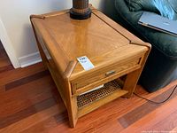 Full view of the side table from an angle showing wooden top, drawer, and bottom woven shelf in a living room setting.
