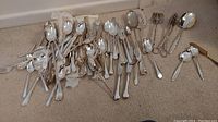 Wide view of assorted silver-plated flatware spread on carpet including forks, spoons, and serving utensils.
