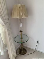 Full view of floor lamp with pleated cloth shade, metal pole, glass table around the pole, and decorative metal base.
