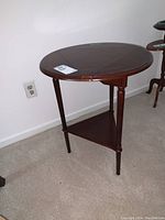 Round wooden drop leaf side table with three tapered legs and triangular lower shelf against a white wall on carpeted floor.