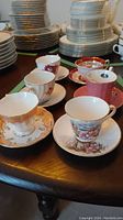 Five assorted vintage teacups and saucers with floral and gold decorative patterns placed on a wooden surface, showing part of a larger collection of stacked china plates in background.
