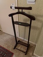 Front view of wooden valet stand showing hanger, trouser bar and base support on carpeted floor.