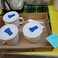 Set of three off-white ceramic mugs with matching lids featuring blue vent flaps, placed in a cardboard box.