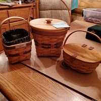 Three Longaberger baskets displayed on a wood surface: one medium with wooden lid, one oval with leather latch, and one small lined basket.