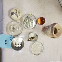Top view of 7 paperweights and a small vase showing various designs and shapes including orange paperweight, clear with bubbles, white oval with text, and two-handled floral vase