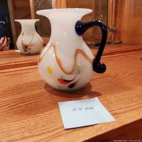 Front and side views of the white opaque glass pitcher with navy blue handle and multicolored abstract accents on a wooden surface in front of a mirror.