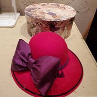 Photo of red felt hat with wide brim and large purple satin bow, placed in front of round hat box featuring vintage collage images.