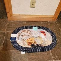 Full view of oval braided rug placed on tile floor, showing painted rooster design at center and size tags attached.