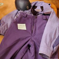 Photo of purple ski pants, light purple ski jacket, and black helmet together on a surface.